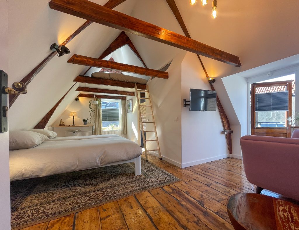 Light room with exposed beams, wooden floor, and two big beds, of which one is accessable via a ladder. There is a sofa and TV in the background as well as access to the roof terrace