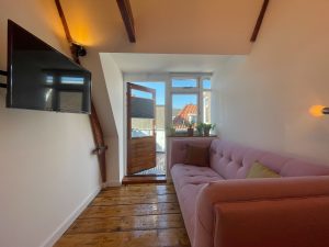 Lichter Raum mit freigelegten Balken, Holzdielen, Sofa, Fernseher und Zugang zur Dachterasse.
